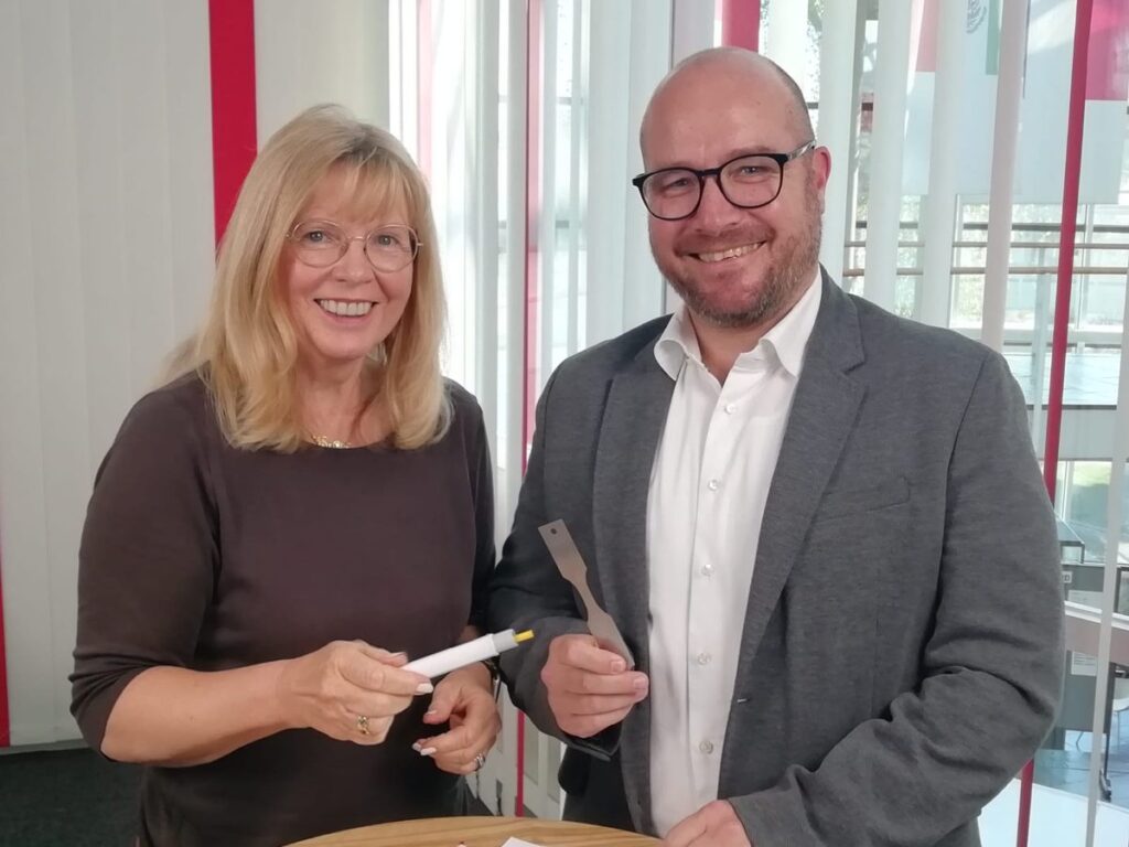Verena Hladik (awikom) und Wolfgang Mörsch (ZwickRoell) zeigen Prüfmuster: einen Autoinjektor und eine Flachzugprobe aus Metall